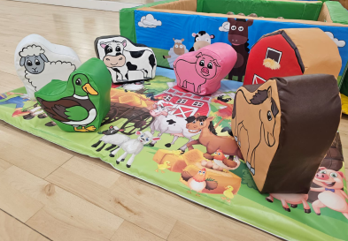 A group soft play items in the shape of farm animals on a farmyard mat with a ball pool in the background.