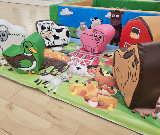 A group soft play items in the shape of farm animals on a farmyard mat with a ball pool in the background.